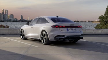 The A5 Saloon e-hybrid stands at an angle in a parking lot on the shore of a lake. Behind it, the skyline of a city can be seen at dusk.