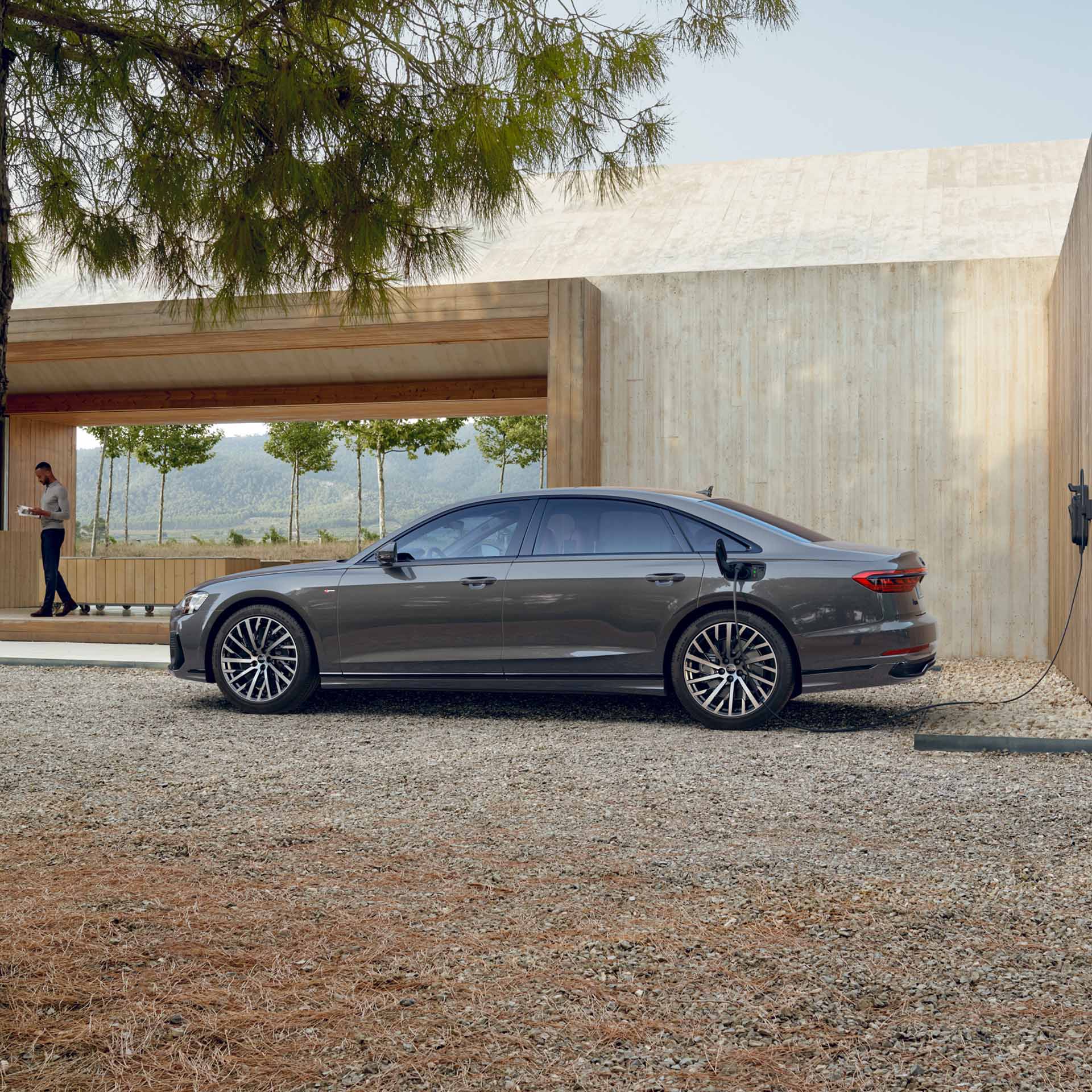 Ein grauer Audi A8 L TFSI e lädt an einer Wandladestation in naturnaher Umgebung mit hellem Holzgebäude. Weit im Hintergrund steht ein farbiger Mann.