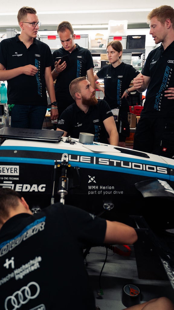 Fünf Personen stehen hinter einem Rennauto und schauen einander an, während eine sechste Person am Auto arbeitet.