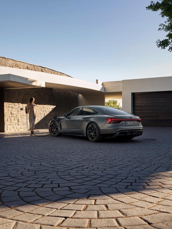 Eine Frau lädt einen grauen Audi RS e-tron GT in einer modernen Einfahrt mit Steinwand und Garage.