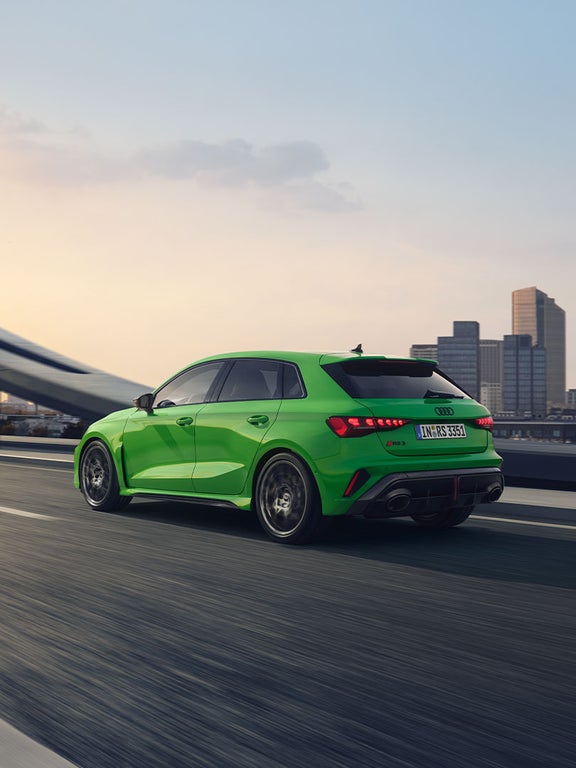 Grüner Audi RS 3 Sportback fährt auf einer Brücke vor städtischer Skyline, schräg von hinten.