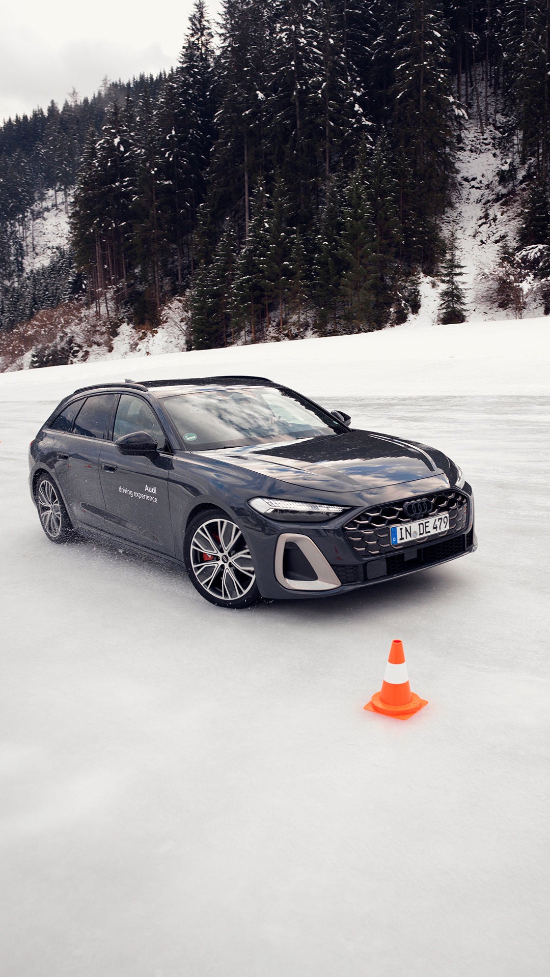 Dunkelgrauer Audi auf vereister Fahrbahn mit Slalomkegeln, umgeben von schneebedecktem Wald im Hintergrund.