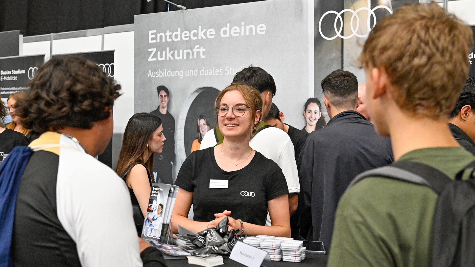 Eine junge Frau im schwarzen T-Shirt mit Audi Logo informiert zwei Interessenten am Messestand.
