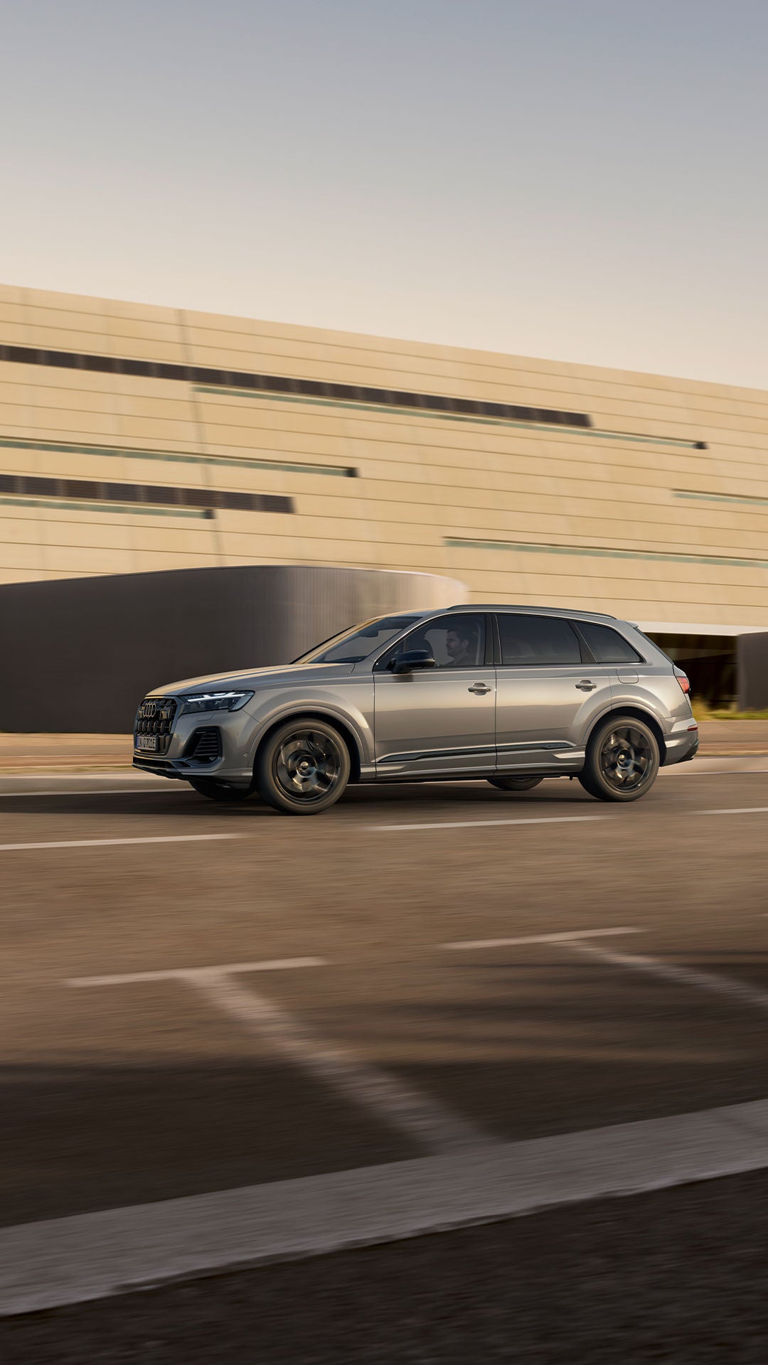 Ein silberner Audi Q7 SUV TFSI e fährt auf Straße an moderner, heller Fassade vorbei, Fahrer sichtbar, dynamische Seitenansicht.
