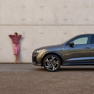Side view of lady in a pink outfit leaning against the wall next to dark grey Audi Q3.
