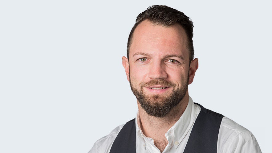 Sebastian Rockstroh: Porträt eines lächelden Mann im mittlerem Alter mit Bart, weißem Hemd mit schwarzer Weste vor grauem Hintergrund.