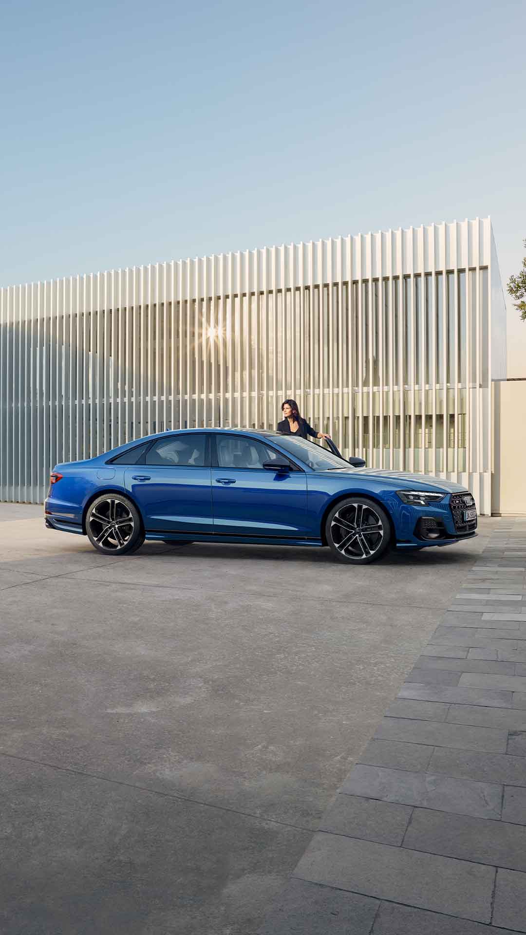 Ein blauer Audi S8 TFSI steht seitlich vor einer Wand mit vertikalen Metalllamellen, dunkelhaarige Frau steigt ein.