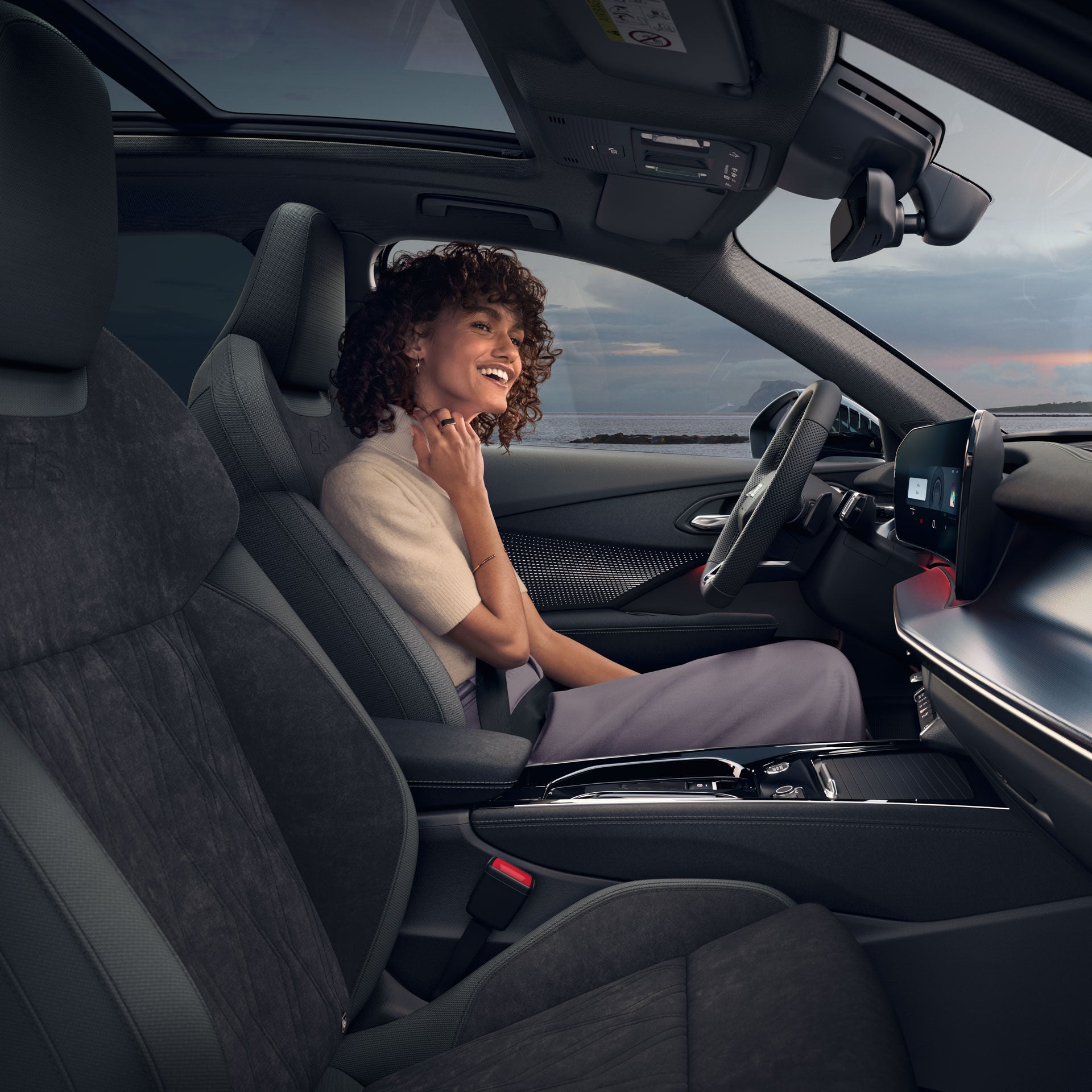 Frau sitzt lächelnd im Fahrersitz eines Audi, Blick auf das Meer durch Fenster und Panoramadach.