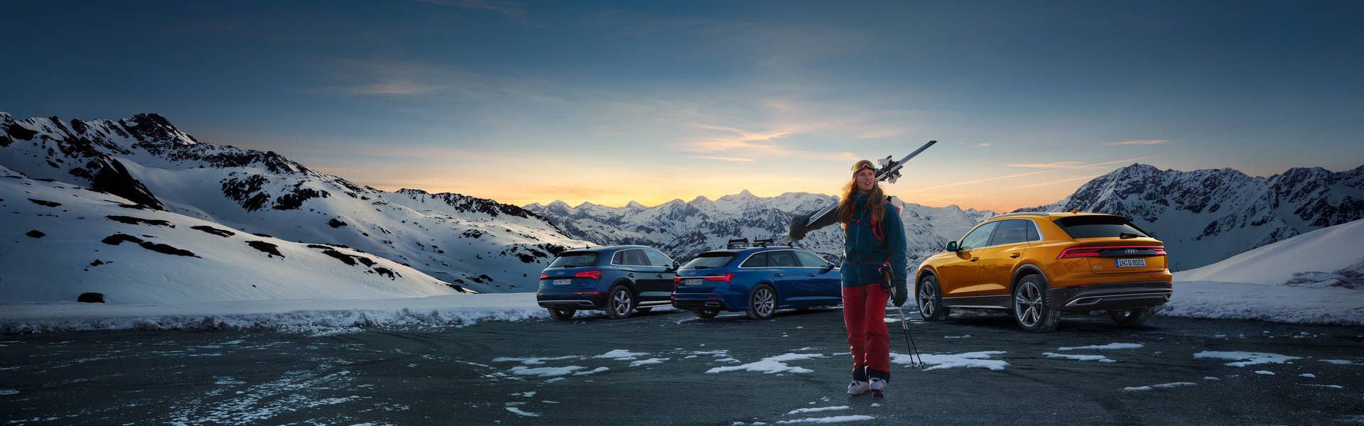 A woman stands with skis in front of a mountainous landscape and various Audi models