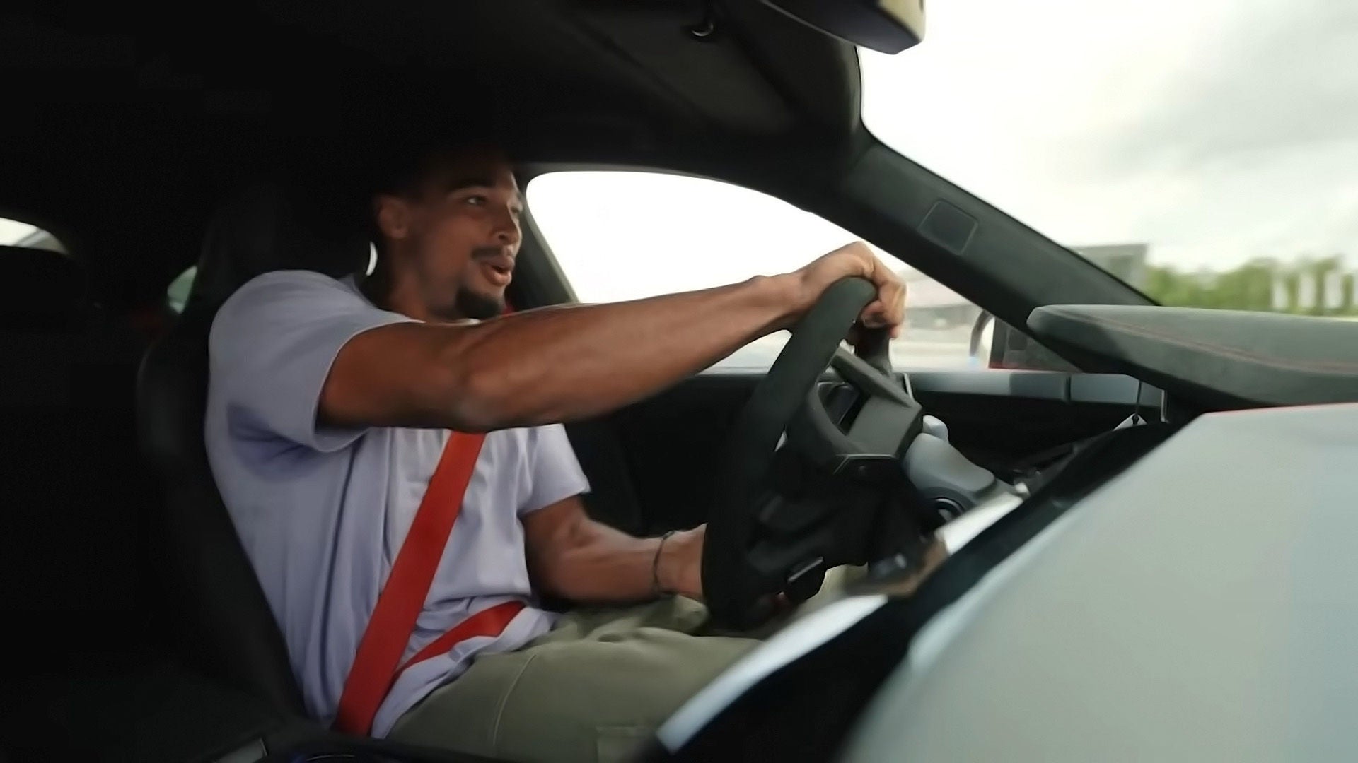 Mann am Steuer eines Sportwagens mit rotem Gurt; Blick nach vorn, dunkler Innenraum, Tageslicht durchs Fenster.