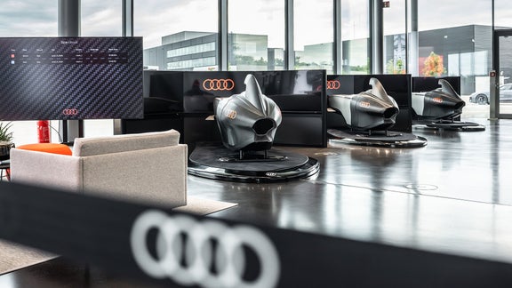 Drei Audi racing Simulator stehen im Showroom; links zeigt ein Bildschirm Zeiten, unscharfe Audi-Ringe sind im Vordergrund.