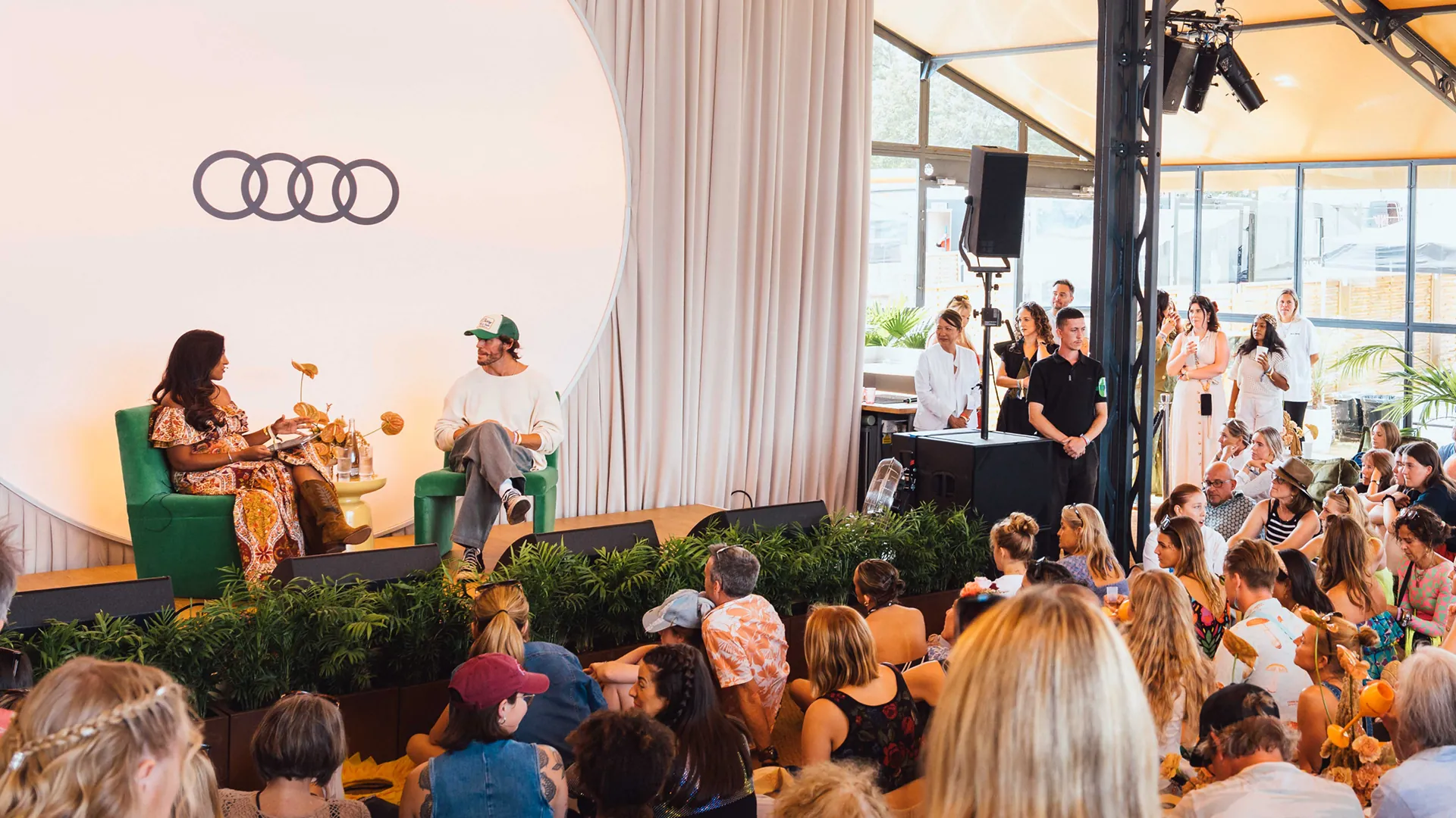 A woman and man chat on green armchairs during an on-stage interview beneath the Audi rings, with a seated audience watching from a plant-lined floor area. A woman and man chat on green armchairs during an on-stage interview beneath the Audi rings, with a seated audience watching from a plant-lined floor area.