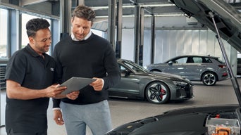 Ein Audi-Verkaufsberater bespricht gemeinsam mit einem Kunden Fahrzeugdetails auf einem Tablet im Audi-Autohaus. Im Hintergrund stehen weitere Audi-Modelle, während im Vordergrund die geöffnete Motorhaube eines Fahrzeugs zu sehen ist.