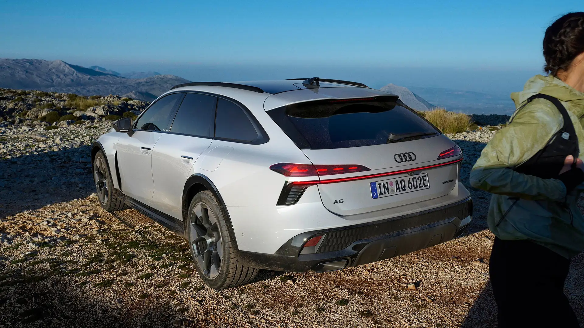 Woman in orange dress and white jacket walking beside a silver Audi RS e-tron GT car on a coastal road.