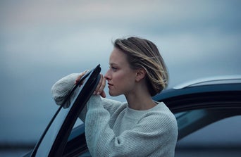 A person in a cozy sweater leans against a car door, gazing thoughtfully at the cloudy sky in a serene outdoor setting.