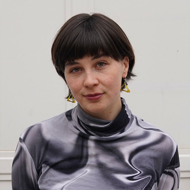 Frau mit kurzem braunem Haar, grauem gemustertem Oberteil und gelben Ohrringen blickt freundlich in die Kamera.