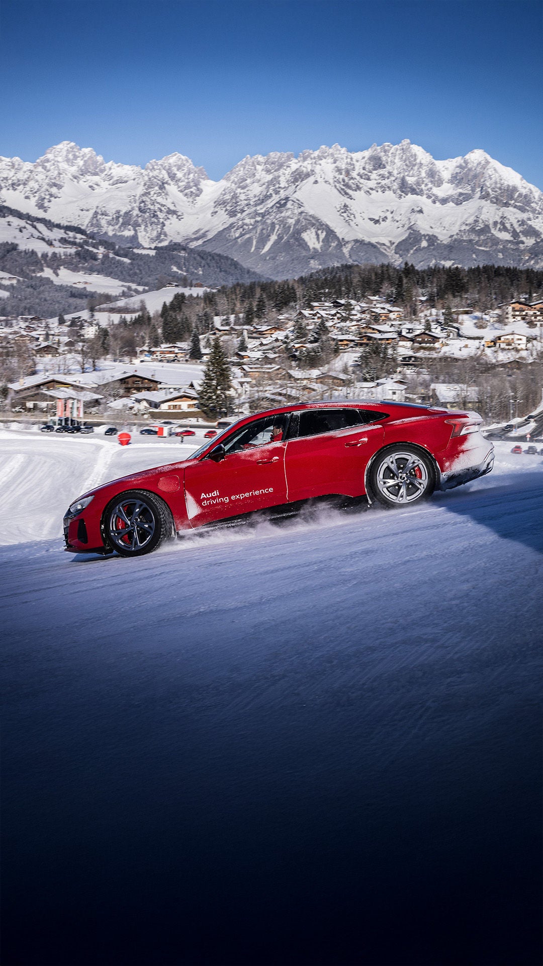 Red car driving up a snow-covered hill; in the background is a snowy Alpine panorama and a winter sports resort with several vehicles.