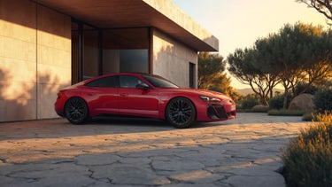 Seitenansicht eines roten Audi e-tron GT quattro, der im Licht eines Sonnenuntergangs auf der gepflasterten Auffahrt vor einem Haus mit moderner Architektur steht.