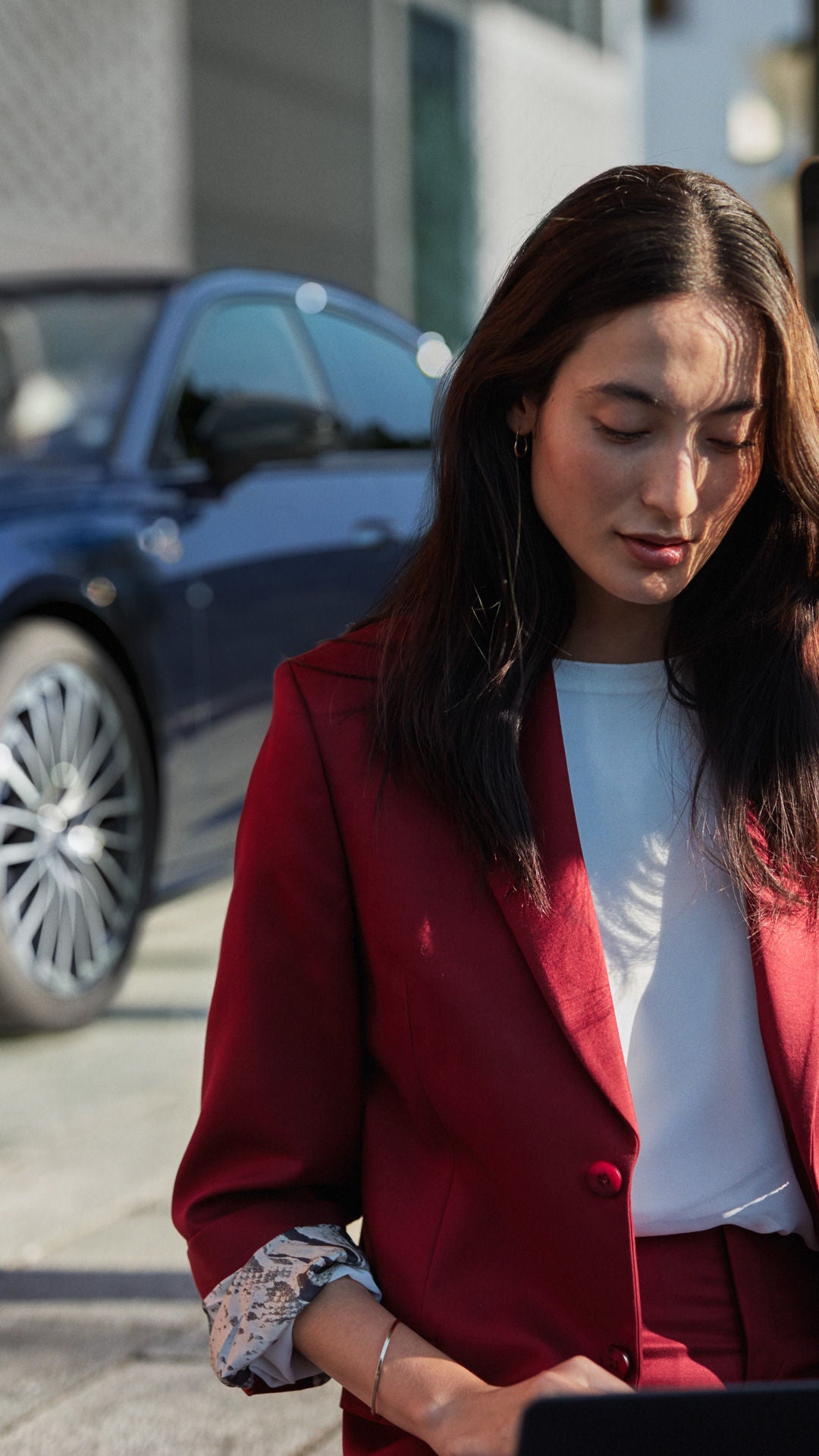 Eine Frau in rotem Hosenanzug sitzt im Freien an einem Laptop. Im Hintergrund steht ein blaues Fahrzeug vor einem Gebäude.