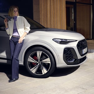 Vista laterale di Audi Q5 SUV e‑hybrid, con una giovane donna vestita alla moda appoggiata al veicolo.