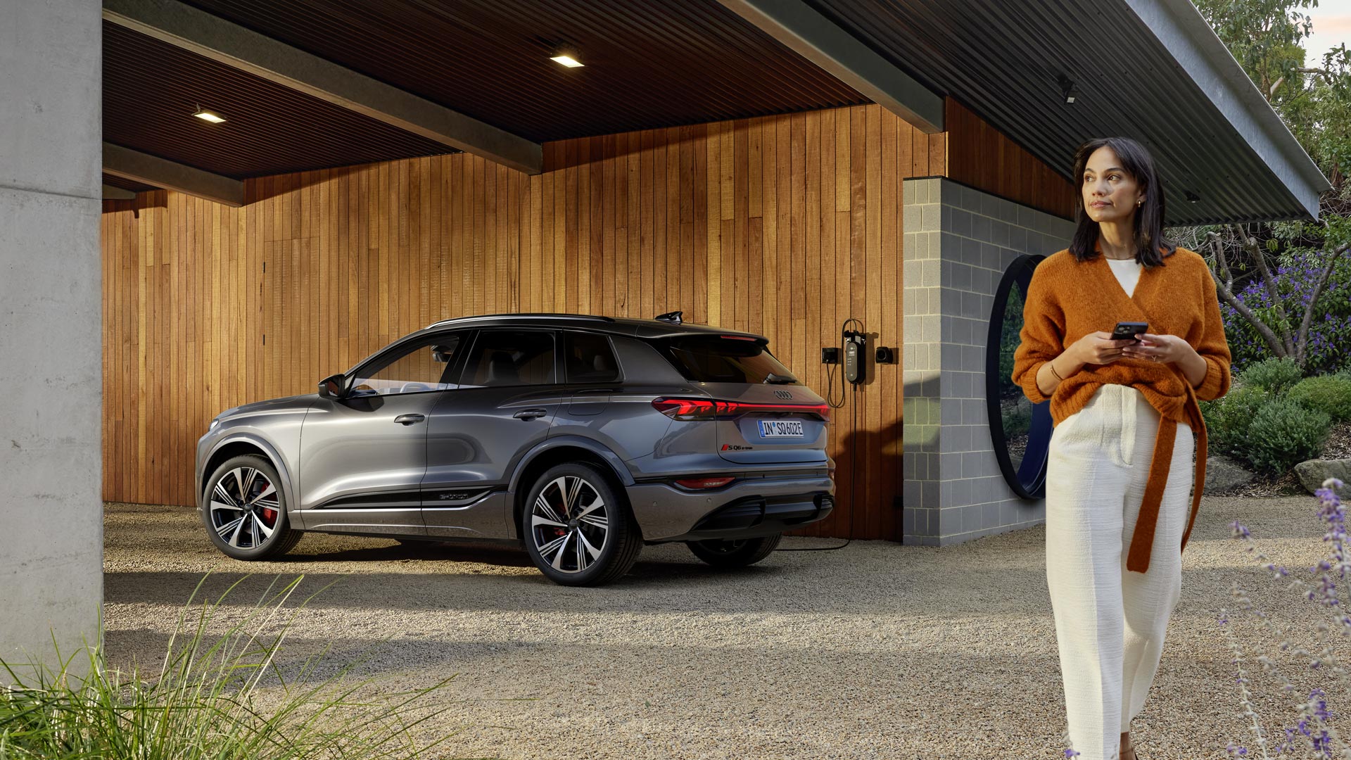 Una mujer está de pie con su smartphone junto a un Audi Q6 e-tron plateado que se está recargando en una estación de recarga doméstica.