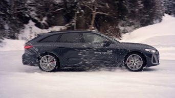 Dunkler Audi S5 Avant mit weißem Schriftzug „Audi driving experience“ fährt auf schneebedeckter Straße, aufgewirbelter Schnee im Fokus.