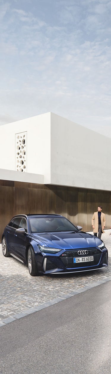 Blauer Audi RS6 Avant parkt vor modernem, weißem Haus mit bronzefarbener Fassade; ein Paar geht Hand in Hand vorbei.