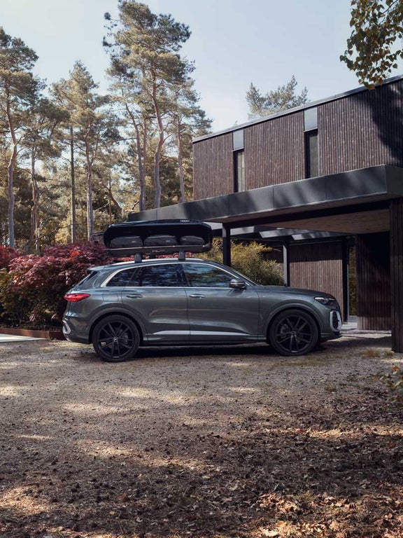 Ein grauer Audi Q5 SUV steht vor einem modernen Holzhaus, mit schwarzer Dachbox und Audi-Taschen darin.