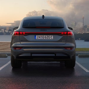 Heckansicht des Q6 Sportback e-tron, welcher auf einem Parkplatz am Meer steht. Im Hintergrund sieht man eine Stadt, welche von Wolken durchzogen ist.