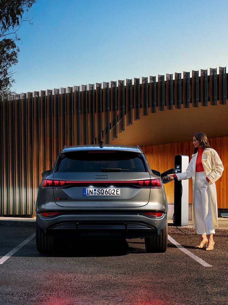 Frau im Trenchcoat lädt grauen Audi SQ6 SUV e-tron an Ladesäule vor moderner Holzfassade, Heckansicht des Fahrzeug.