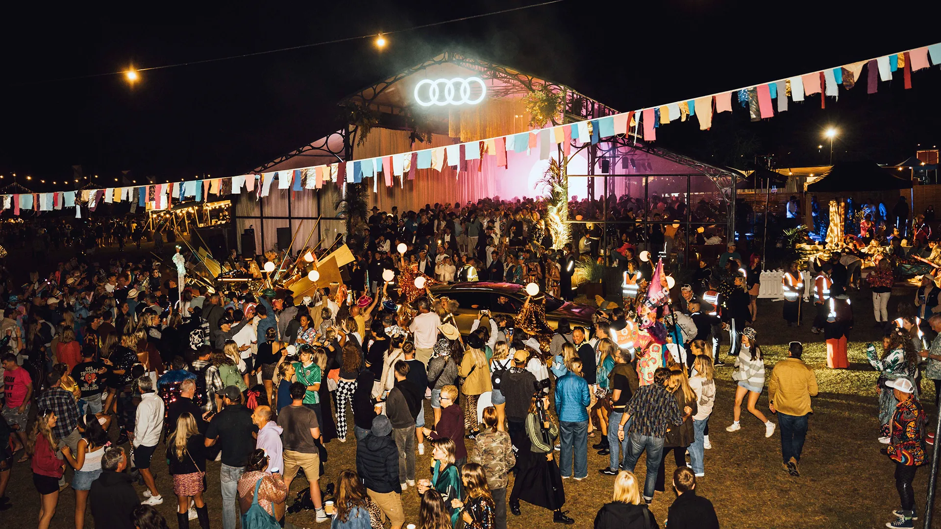 A large nighttime crowd gathers beneath bunting and the illuminated Audi rings outside the festival pavilion, with performers and a car visible amid the celebration. A large nighttime crowd gathers beneath bunting and the illuminated Audi rings outside the festival pavilion, with performers and a car visible amid the celebration.