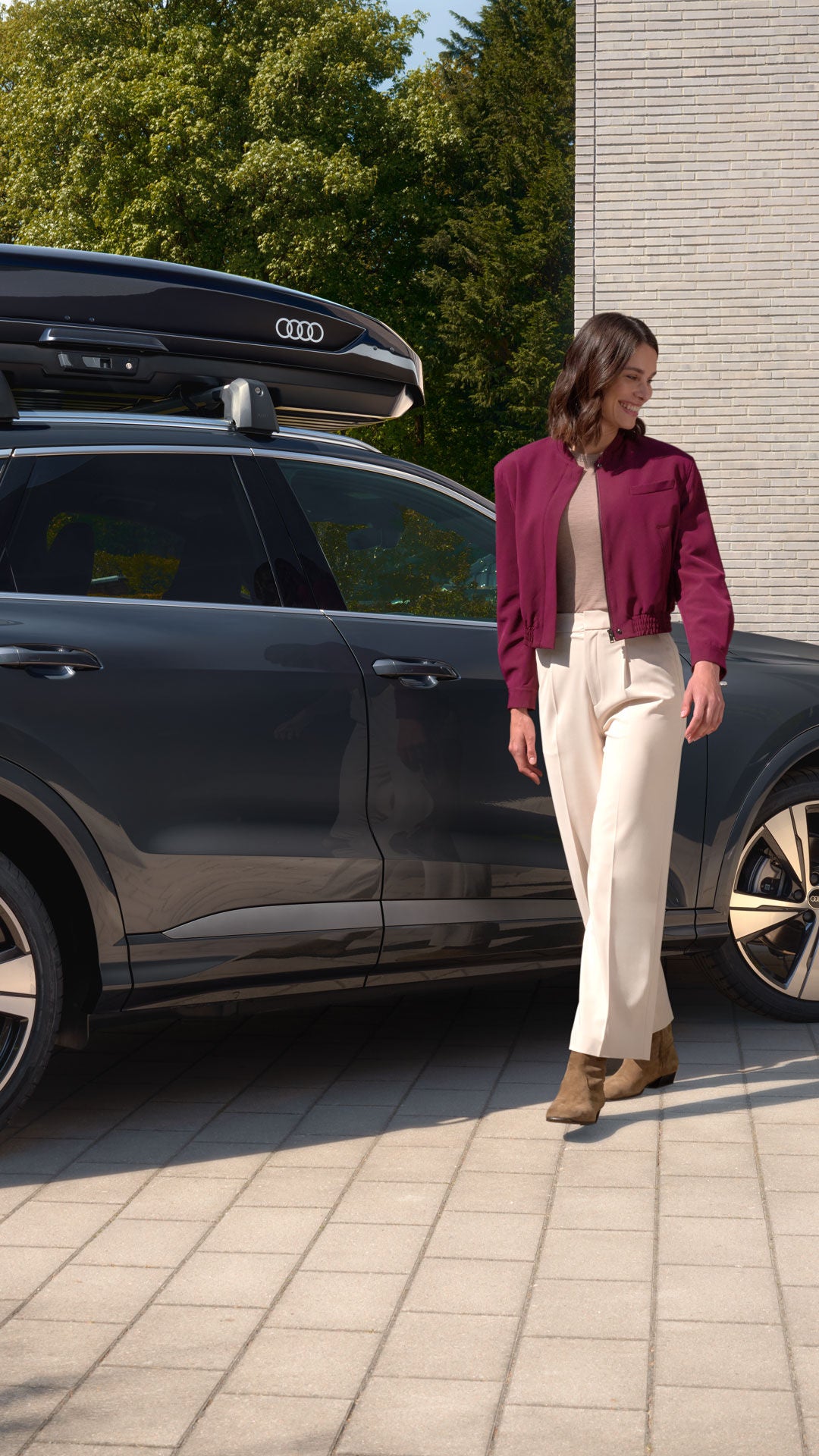 Grauer Audi SUV mit Dachbox vor Wohnhaus; Frau geht mit Hund, Mann steht im Eingangsbereich.