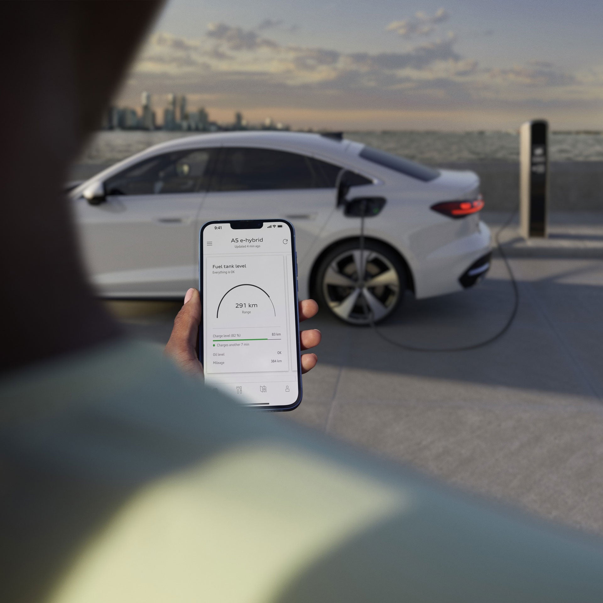 Handybildschirm zeigt den Ladestand des im Hintergrund an der Ladesäule stehenden, weißen Audi an