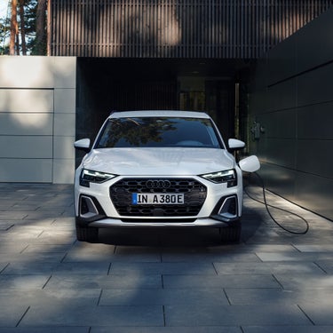 Frontansicht eines weißen Audi A3 allstreet TFSI e, der mit einem Ladekabel an einer Wallbox in einer Einfahrt aufgeladen wird.