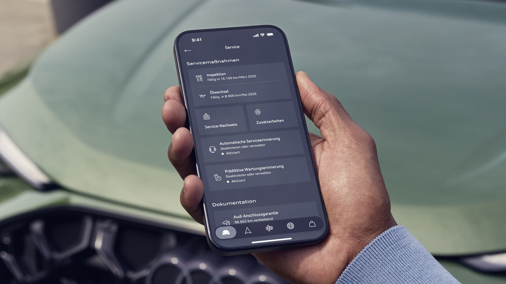 Die Hand eines Mannes ragt von unten ins Bild. Er hält ein Smartphone in der Hand, auf dessen Display Service-Funktionen angeboten werden. Im Hintergrund unscharf ein grüner Audi Q5.