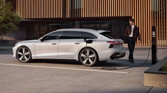 The A6 Avant e-hybrid is parked sideways in front of the entrance to a large building and is being charged via a power cable. A middle-aged man is standing at the rear of the vehicle.