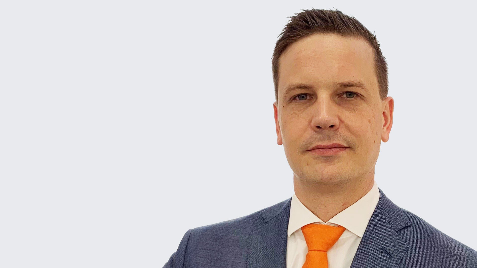 Daniel Schwarz: Porträt eines Mannes im blauen Anzug mit weißem Hemd und orangefarbener Krawatte vor hellgrauem Hintergrund.