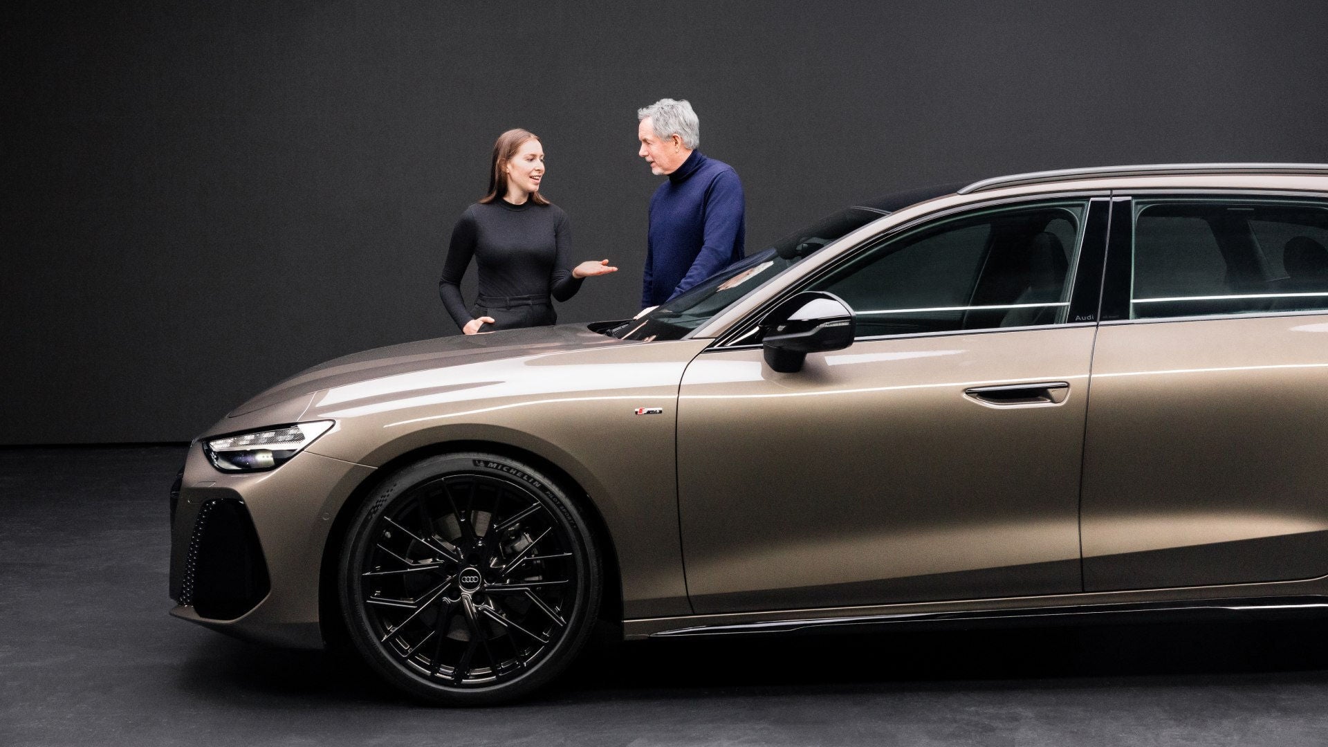 Zwei Personen im Gespräch neben einem goldenen Audi A6 Avant mit schwarzem Felgendesign.