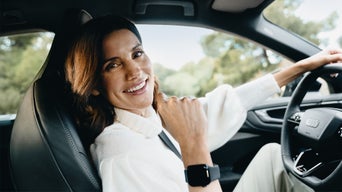 Eine Frau sitzt in einem Auto und zeigt lächelnd ihre Smartwatch am rechten Handgelenkt Richtung Kamera. 