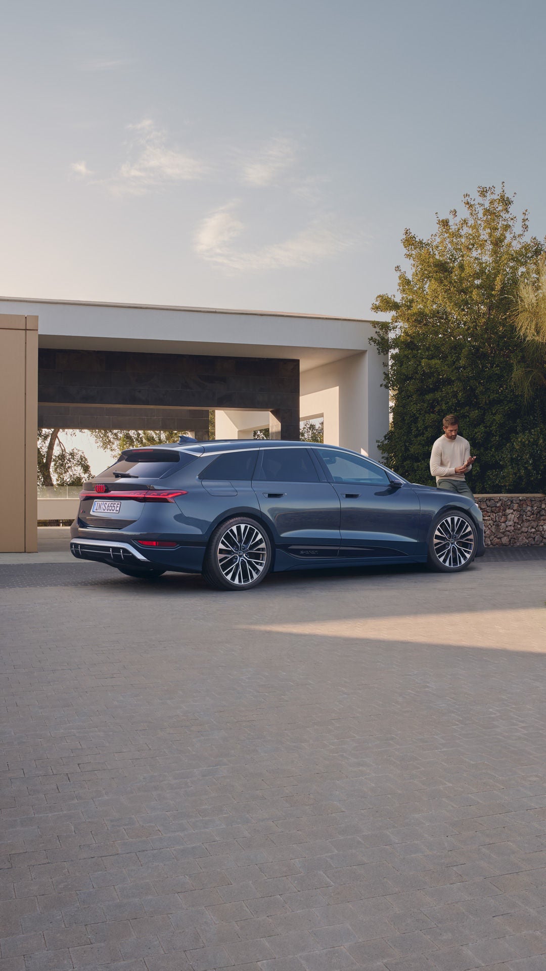 Ein blauer Audi S6 Avant e-tron in seitlicher Heckansicht vor einem modernen Wohnhaus. Im Hintergrund lehnt ein Mann an einer Steinmauer und blickt auf sein Smartphone.