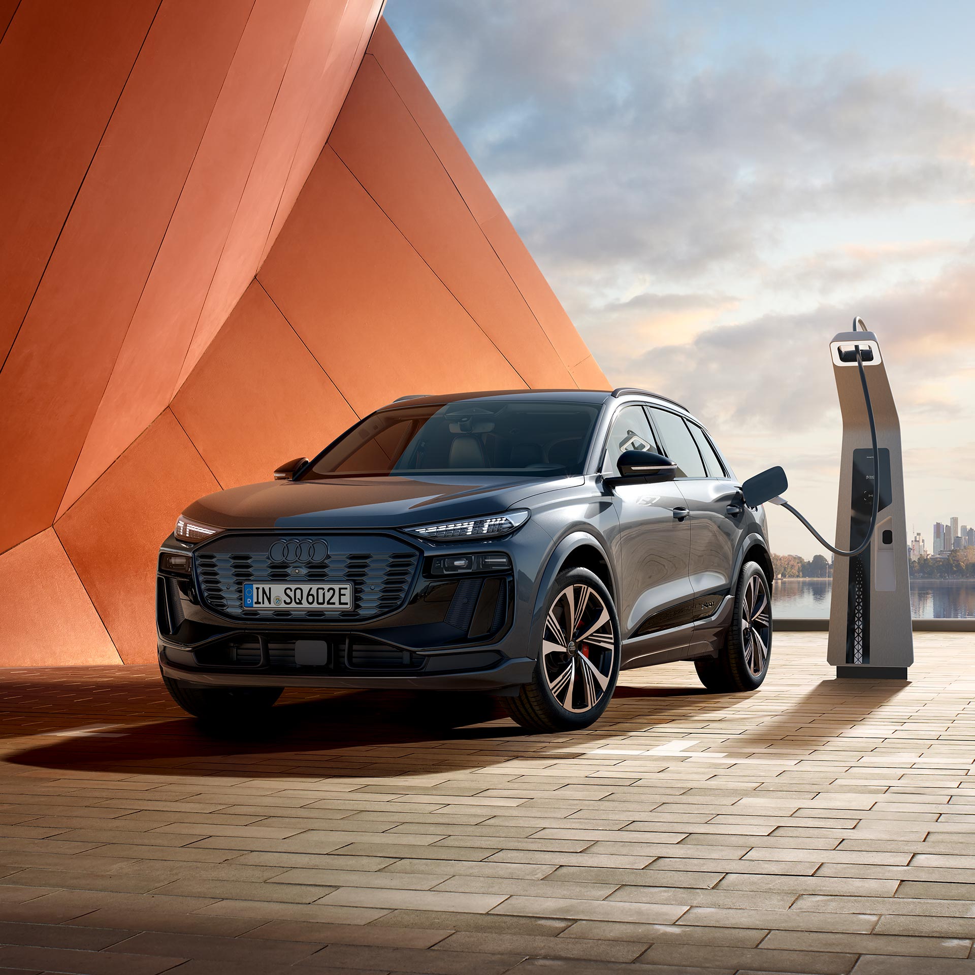 Ein grauer Audi SQ6 SUV e-tron lädt an Ladesäule vor oranger Architektur, im Hintergrund Wasserfläche und Skyline.