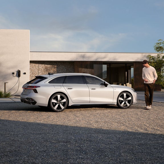 Der A6 Avant e-hybrid steht im Seitenprofil vor einem modernen Haus und ist an eine Ladestation angeschlossen. Ein Mann mittleren Alters steht vor dem Fahrzeug.