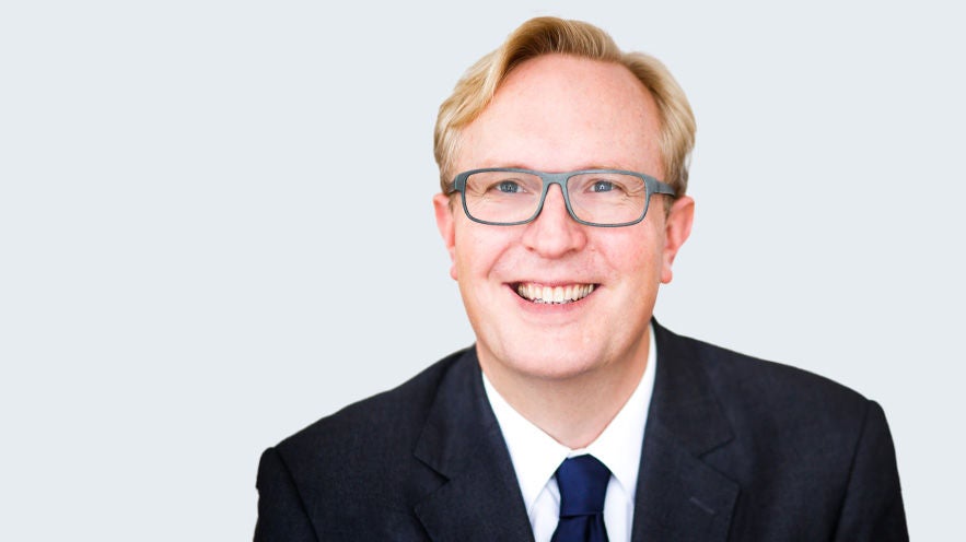 Dr. Lars Siebert: Porträt eines älteren, lächelnden Mann mit hellblondem Haar und grauer Brille im Anzug vor grauem Hintergrund.