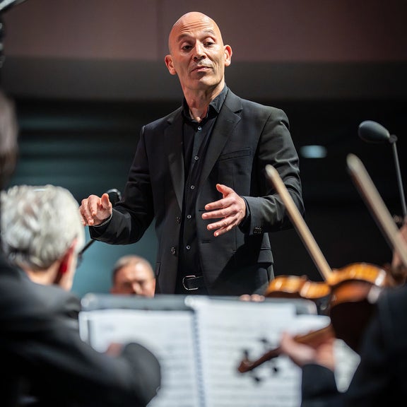 Porträt eines Dirigenten im schwarzen Anzug, Hände erhoben, mit fokussiertem Blick während einer Orchesteraufführung.