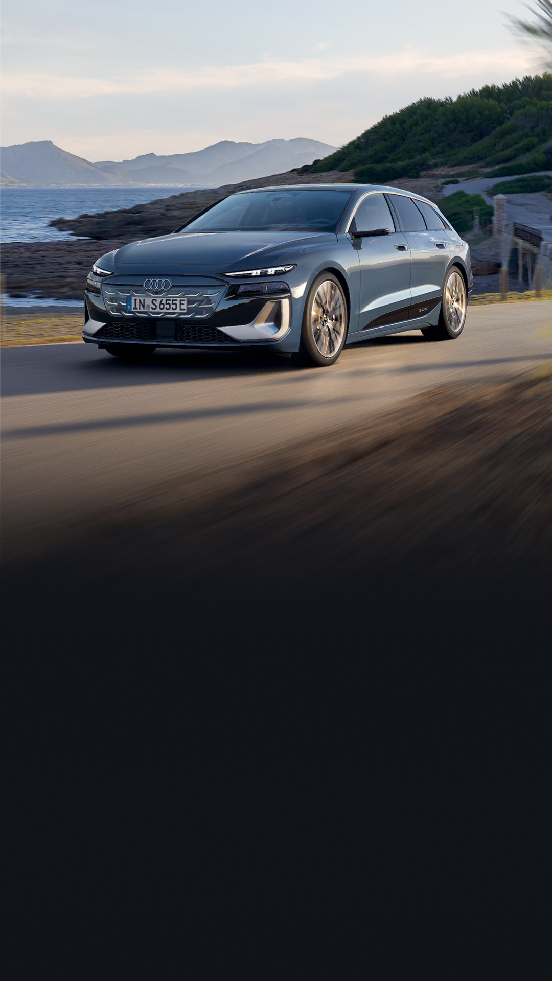 The S6 Avant e-tron seen from an angle as it drives along a coastal road. Mountains can be seen in the background.