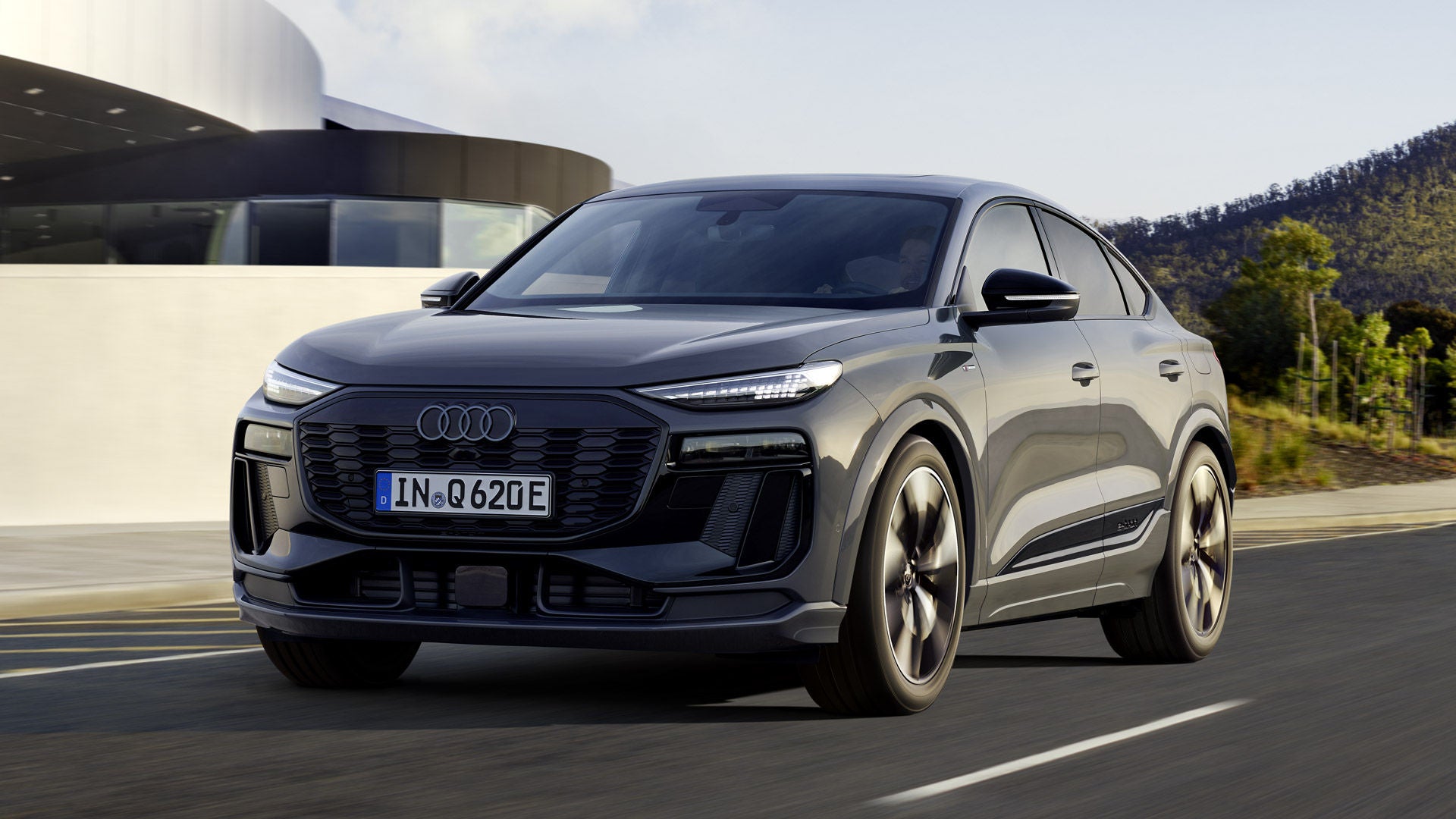 Schräges Frontprofil des Q6 Sportback e-tron, welcher auf einer Straße neben einem modernen Haus fährt.