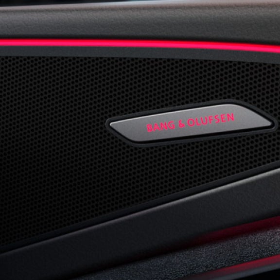 Close-up of a sleek car audio speaker grille featuring the Bang & Olufsen logo illuminated by subtle red backlighting.
