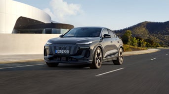 A sleek black Audi Q6 Sportback e-tron drives on a scenic road, modern architecture and hills visible in the background under a clear sky.