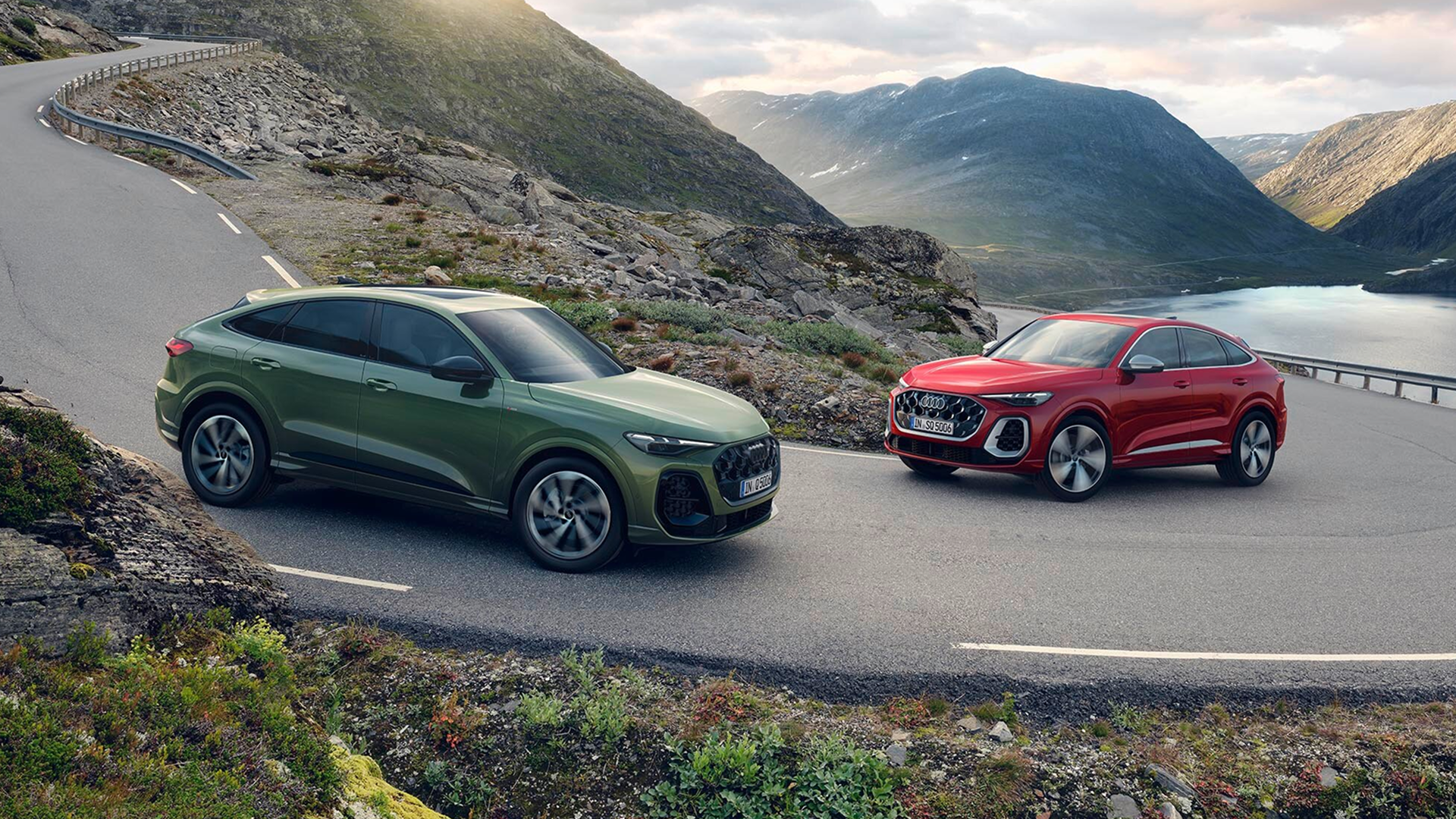 Two Audi SUVs, one green and one red, crossing on a winding mountain road with scenic hills and a lake in the background.