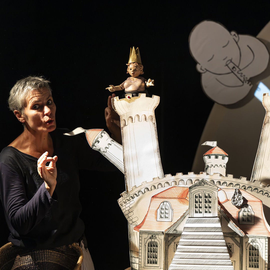 Frau mit kurzen grauen Haaren steht neben einem gezeichneten Schloss; oben eine Puppenfigur mit Krone vor schwarzem Hintergrund.